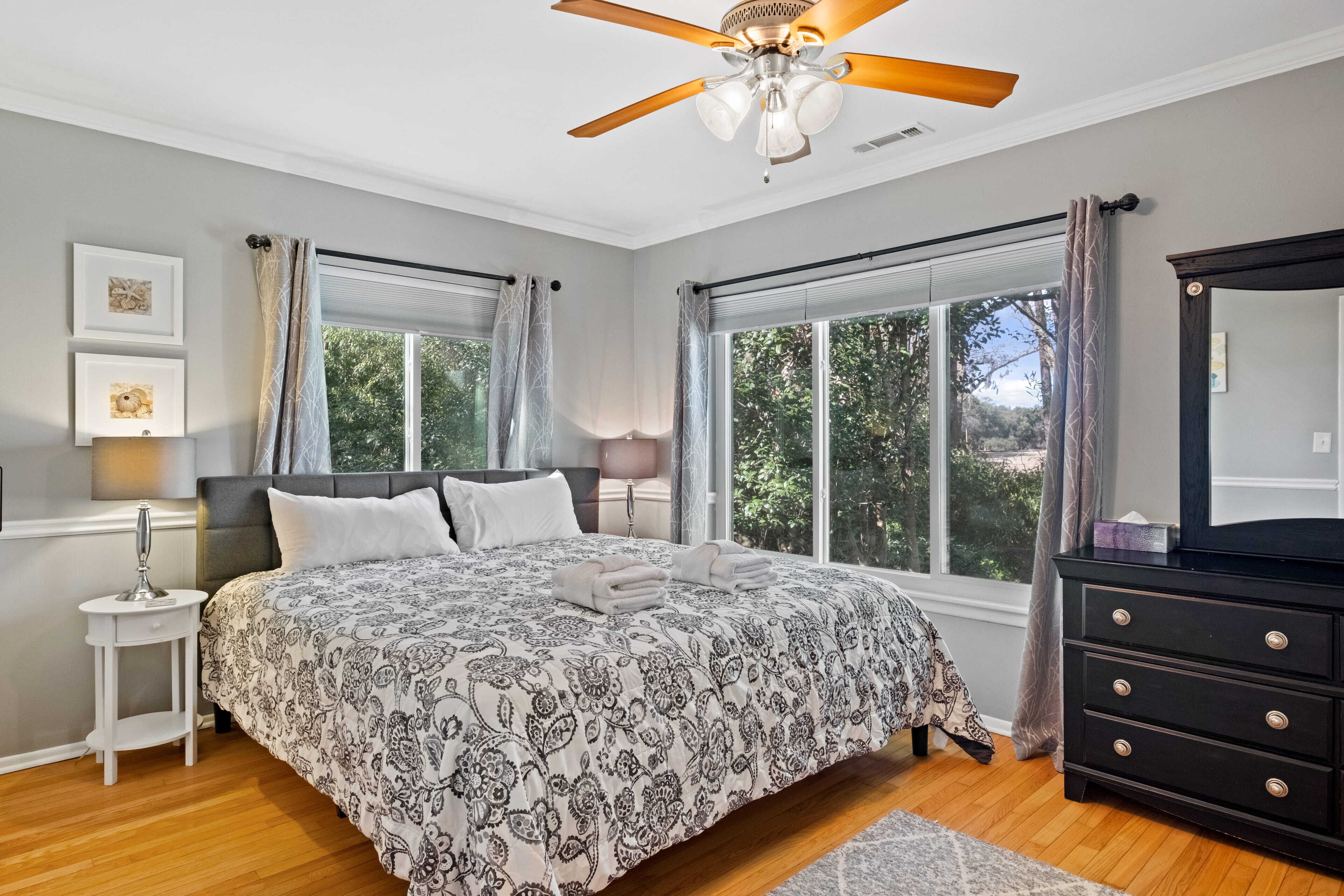 A bedroom with a large bed, a ceiling fan, a dresser, and a window with curtains.
