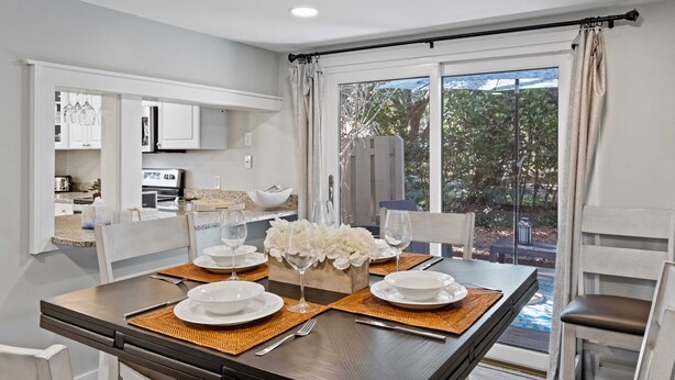 Comedor con una mesa dispuesta para cuatro, sillas blancas y vistas al exterior a través de grandes ventanales.