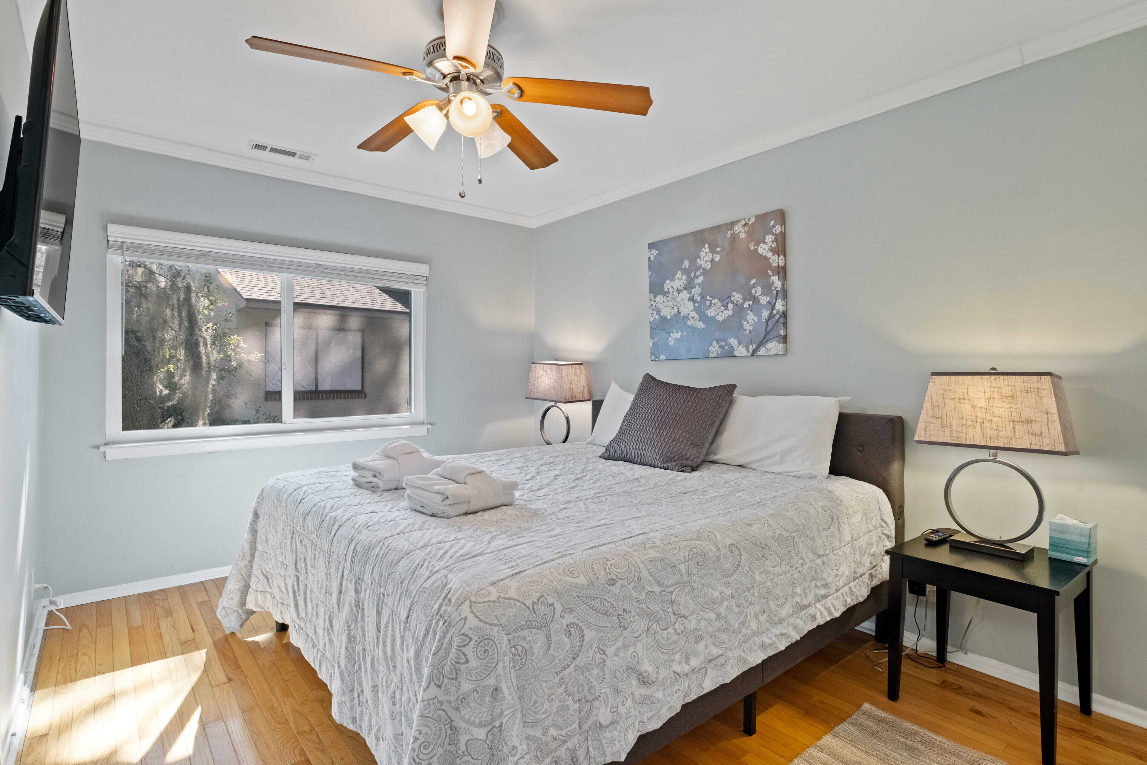 A bedroom with a bed, a ceiling fan, a window, a nightstand, and a wall-mounted TV.