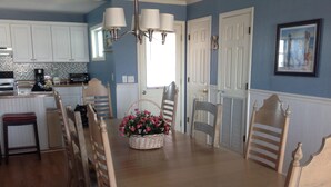 A dining area with a wooden table, six chairs, a basket of flowers, and a kitchen in the background. -  @ Ocean Front Guest Friendly Home "An Emerald On The Beach" (Sunset Beach)