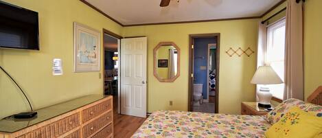 A bedroom with a bed, dresser, ceiling fan, and a television on the wall.