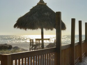 A wooden deck with a thatched umbrella and chairs overlooking the ocean at sunset.