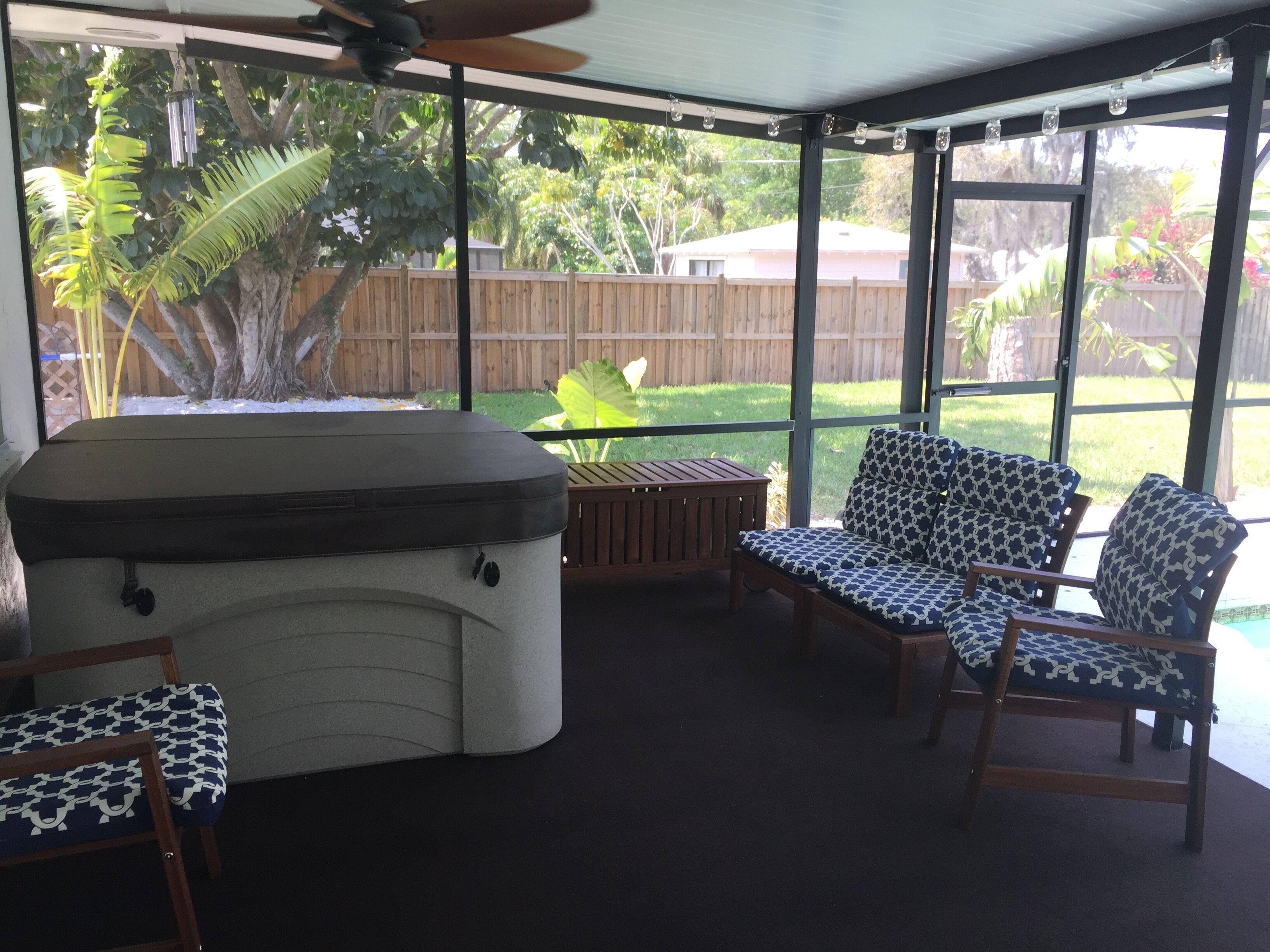 A covered patio area with a hot tub, two patterned chairs, and a wooden table.