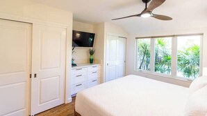 A bedroom with a bed, a ceiling fan, a TV, and a window with a view of greenery.