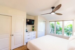 A bedroom with a bed, a ceiling fan, a TV, and a window with a view of greenery.