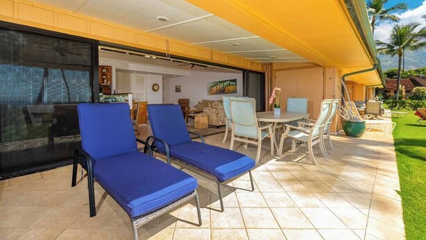 A covered outdoor seating area with lounge chairs and a table, a glass door leading to an interior space, and a view of a lawn with palm trees.