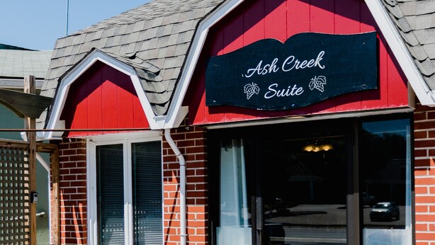 A brick building with a red and white facade, a sign reading "Ash Creek Suite", and outdoor seating with tables and chairs. - Ash Creek Suite - In Downtown Historic Hermann (Hermann)