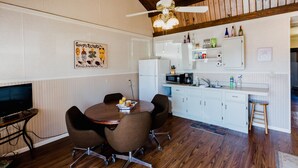 A compact kitchen with white cabinets, a microwave, and a refrigerator. There is a dining table with four chairs and a ceiling fan above. - Ash Creek Suite - In Downtown Historic Hermann (Hermann)