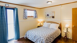 A bedroom with a bed, two bedside tables with lamps, a window with patterned curtains, and a wall-mounted artwork. - Ash Creek Suite - In Downtown Historic Hermann (Hermann)