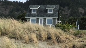 A house with a sloped roof, two windows, and a door, surrounded by tall grass and a forested hill in the background. - Oceanfront Bungalow With Beach Access On The Oregon Coast (Cloverdale)