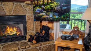 A stone fireplace with a TV mounted above it, a bear-themed table with a game controller, and a view of greenery through a window.