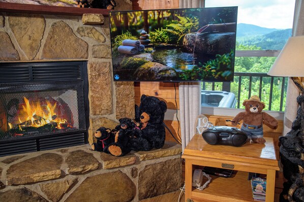 A stone fireplace with a TV mounted above it, a bear-themed table with a game controller, and a view of greenery through a window.