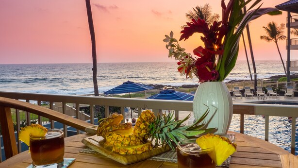 A wooden deck with a table set for two, featuring a tropical drink, a pineapple, and a vase of flowers. The deck overlooks a beach with umbrellas and palm trees, and the ocean is visible in the background.