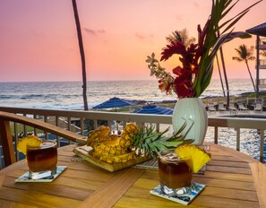 Een houten terras met een tafel voor twee, met een tropische cocktail, een ananas en een bloevas. Het terras kijkt uit over een strand met parasols en palmbomen, en de oceaan is in de achtergrond zichtbaar.