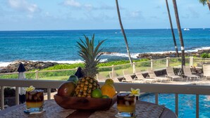 A tropical setting with a view of the ocean, a poolside table with a fruit bowl and drinks, and lounge chairs.