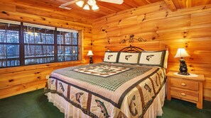 A cozy log cabin bedroom with a large bed, two bedside lamps, a nightstand, and a window with a view of trees. - Summer Specials, 4 min to Parkway, Mountain Views, Hot Tub, Game Room. (Pigeon Forge)