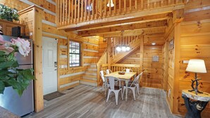 Interior de una cabaña de madera con una mesa de comedor, sillas, un refrigerador y una escalera.