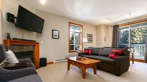 A living room with a fireplace, a flat-screen TV mounted on the wall, a sofa with red and black pillows, and a large window with a view of trees.