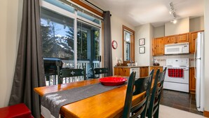 A dining area with a wooden table, chairs, and a red bowl. A kitchen with wooden cabinets, a microwave, and a refrigerator is visible in the background.