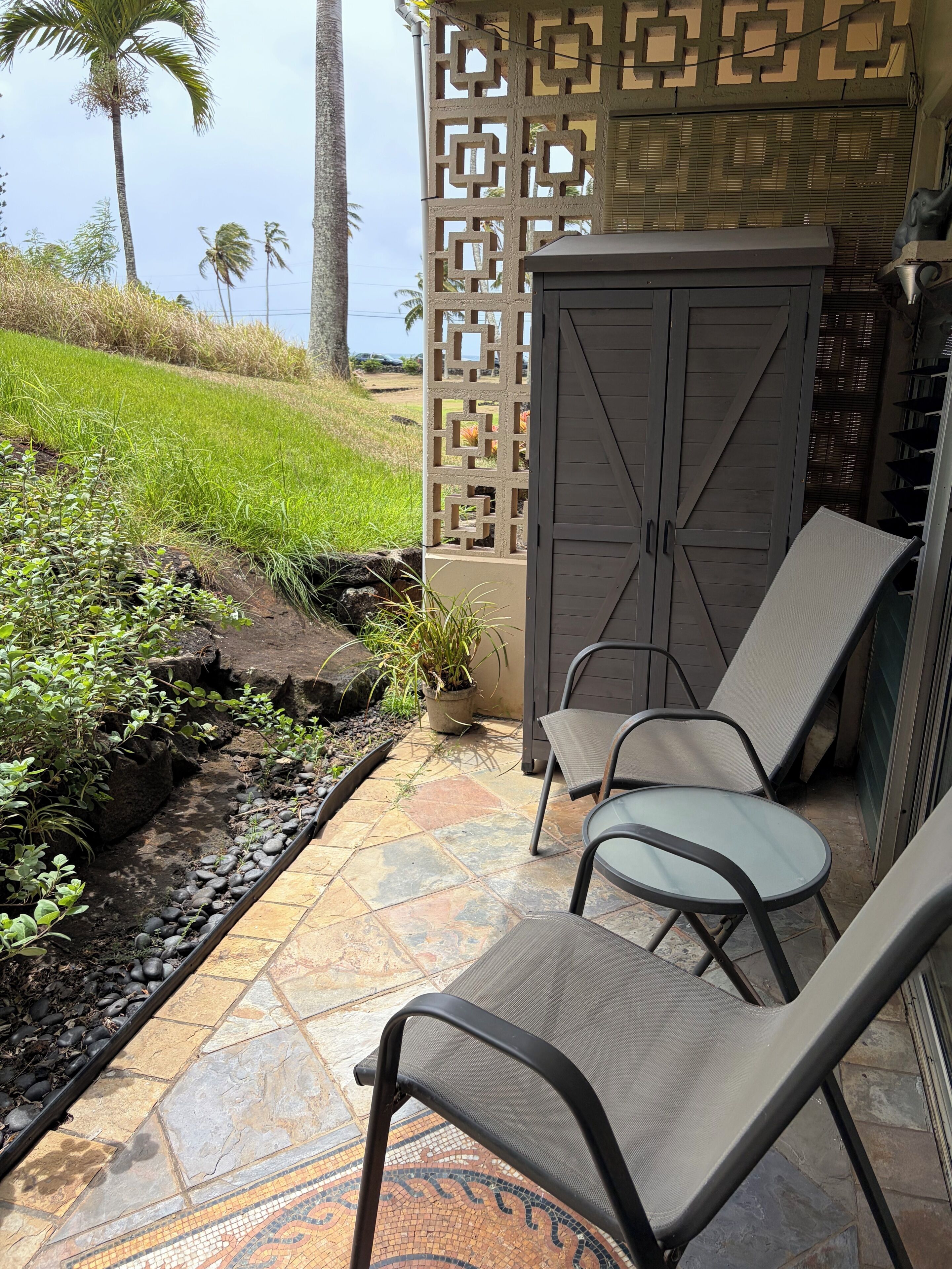 An outdoor seating area with two chairs and a cabinet, surrounded by a garden with palm trees and a decorative wall.