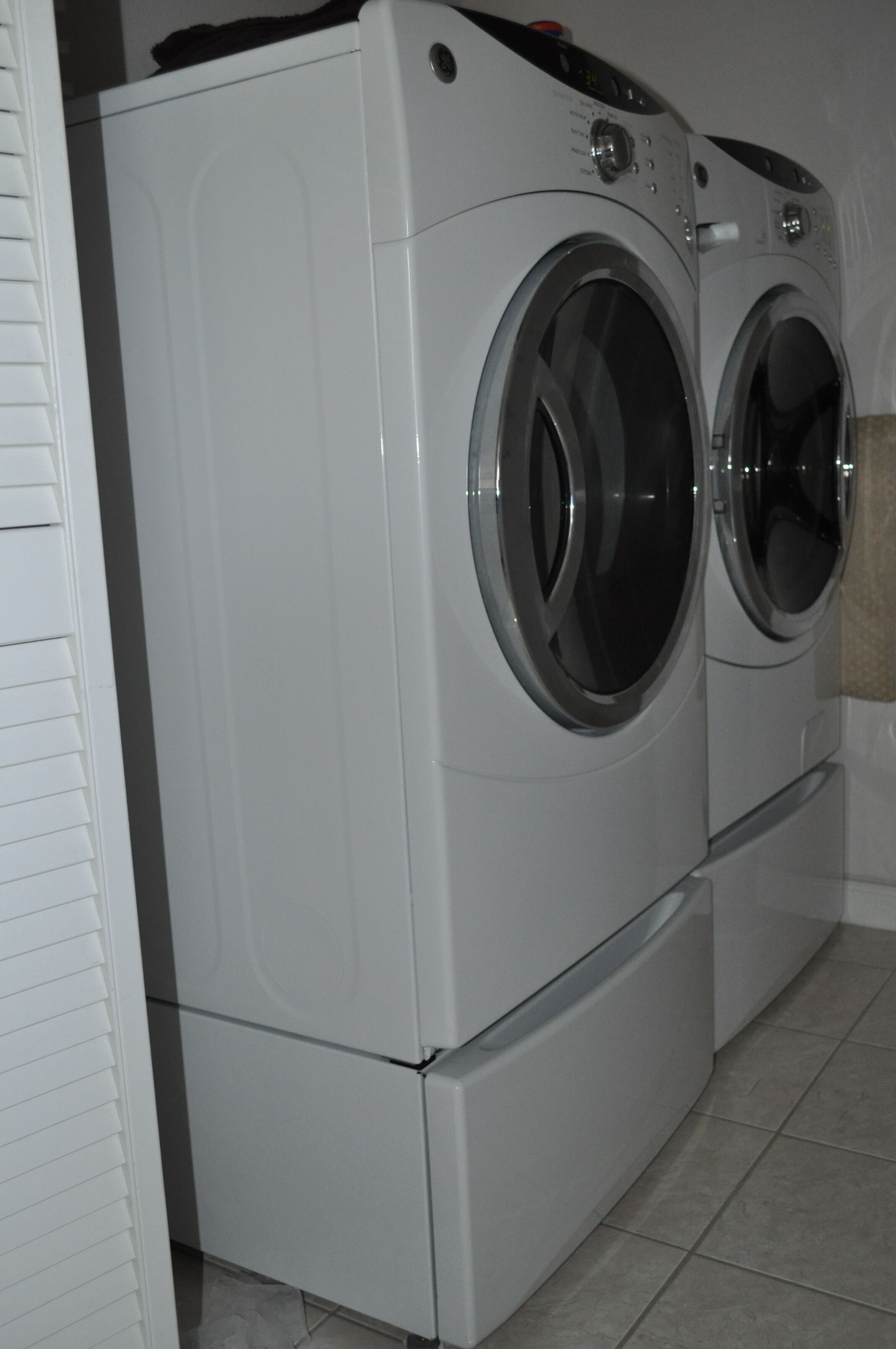 Vue de face d’un appareil combiné lave-linge et sèche-linge compact, blanc et à deux portes, installé dans une salle de bain dont le sol est carrelé.