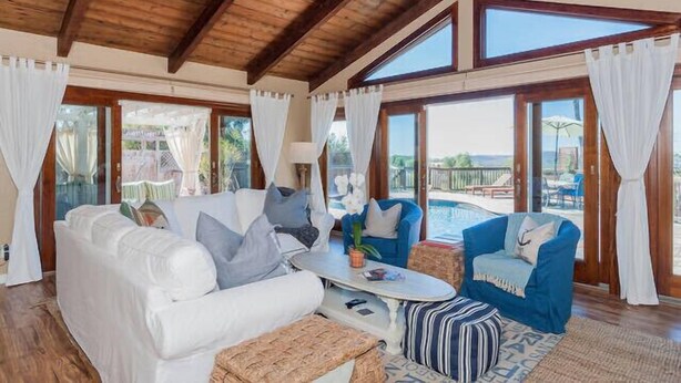 A spacious living room with a white sofa, blue armchair, and a coffee table. Large windows provide a view of the outdoors.
