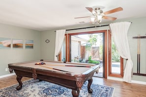 A pool table in a room with a ceiling fan, a view to the outside, and a set of pool cues on the wall.