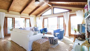 A spacious living room with a high wooden ceiling, a white sofa, blue armchairs, and a large window providing natural light.
