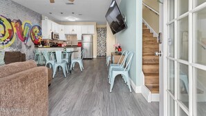 A modern kitchen with white cabinets, a microwave, and a refrigerator. There is a dining area with light blue chairs and a table. A ceiling fan is visible, and a flat-screen TV is mounted on the wall. - Blue Beach House, GAME ROOM, Sleep 8, Swim Pool, Gated, Casinos (Gulfport)
