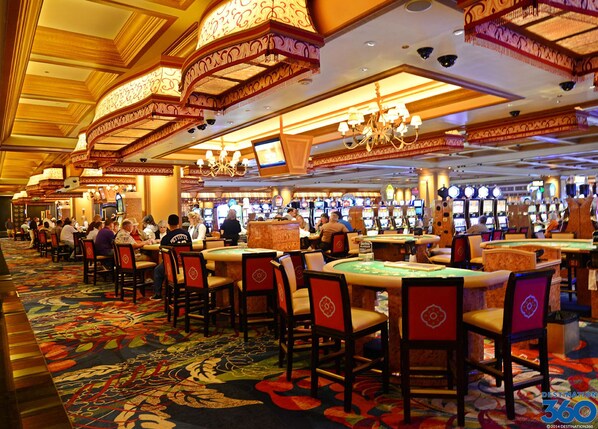 A casino with multiple gaming tables and chairs, a patterned carpet, and ornate ceiling decorations.