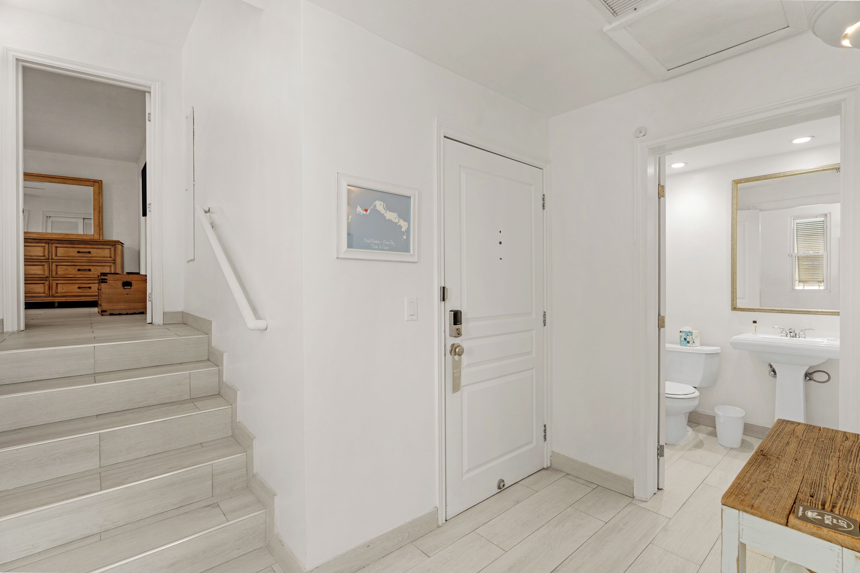 Un baño moderno y limpio, con lavamanos, inodoro y zona de ducha blancos. Hay una mesita de madera con un espejo encima.