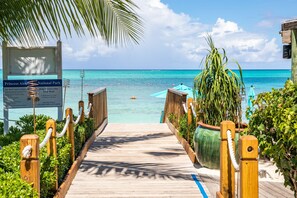 A wooden boardwalk leading to a beach with clear turquoise water, a signboard, and a palm tree.