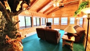 A cozy cabin interior with a wooden ceiling, large windows, a television, a sofa, and a lamp.