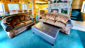A cozy log cabin interior with two patterned sofas, a coffee table, a stone fireplace, and a dining area with wooden furniture.