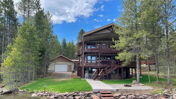 A multi-story log cabin with a dock extending into a pond, surrounded by trees.