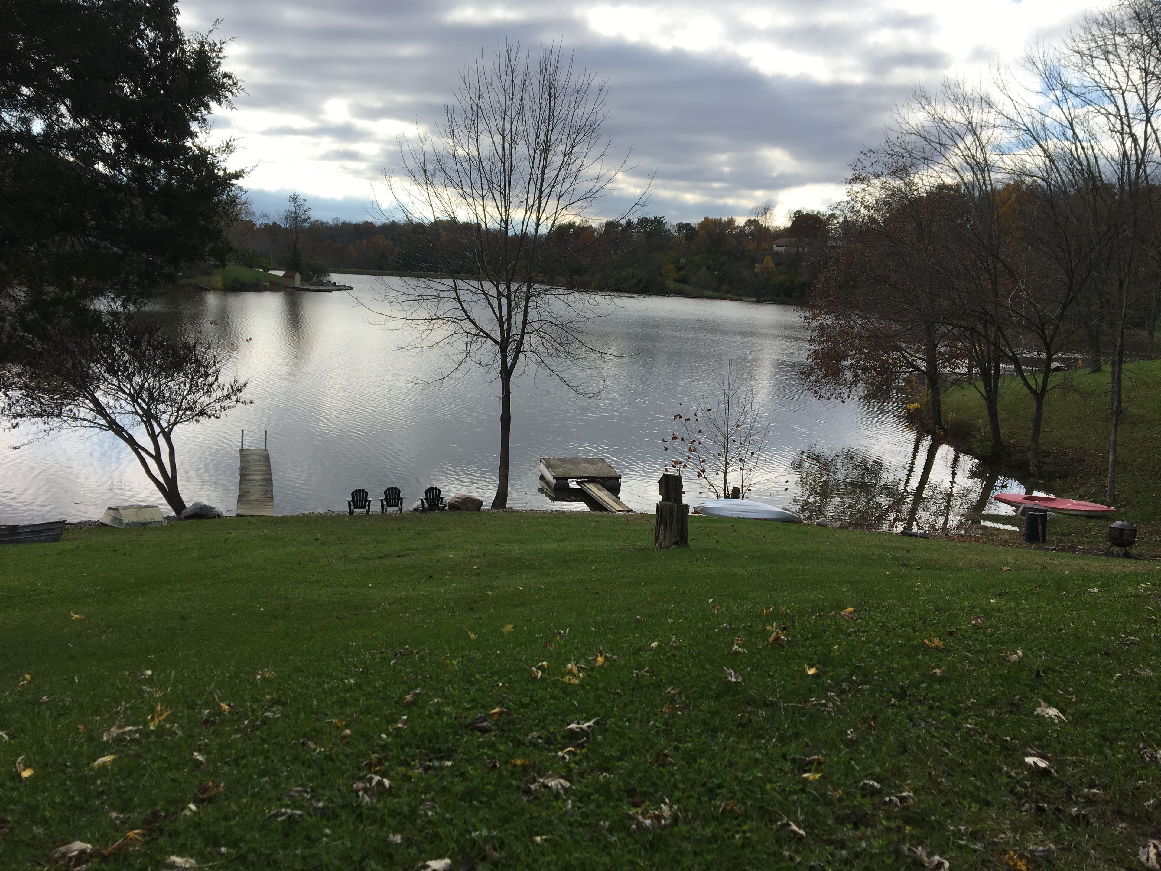 A lakeside park with a grassy area, a wooden dock, and a row of chairs facing the water.