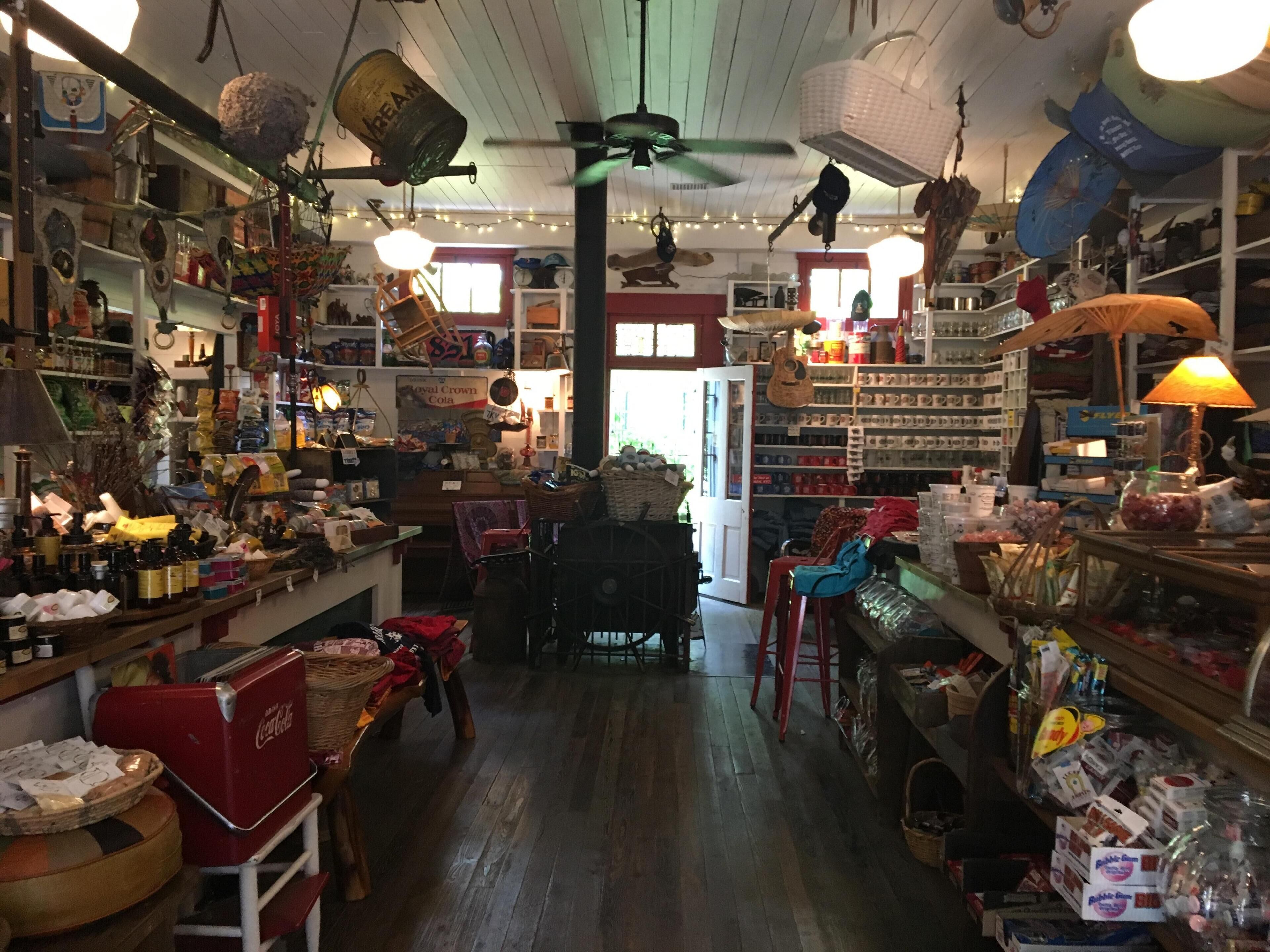 Una tienda desordenada con estantes llenos de diversos artículos, un refrigerador rojo de Coca-Cola y un ventilador de techo.