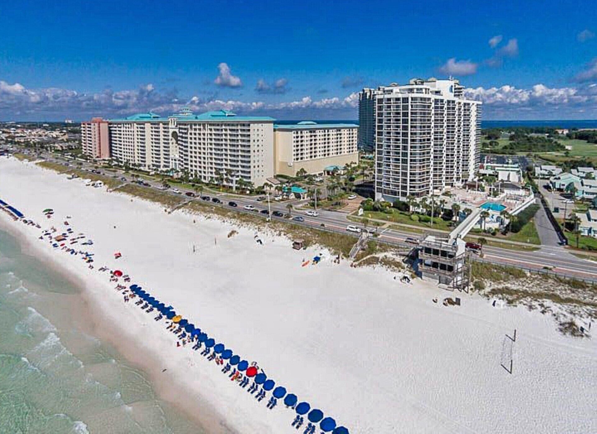 A coastal hotel with multiple high-rise buildings, a beach with umbrellas, and a clear view of the ocean.