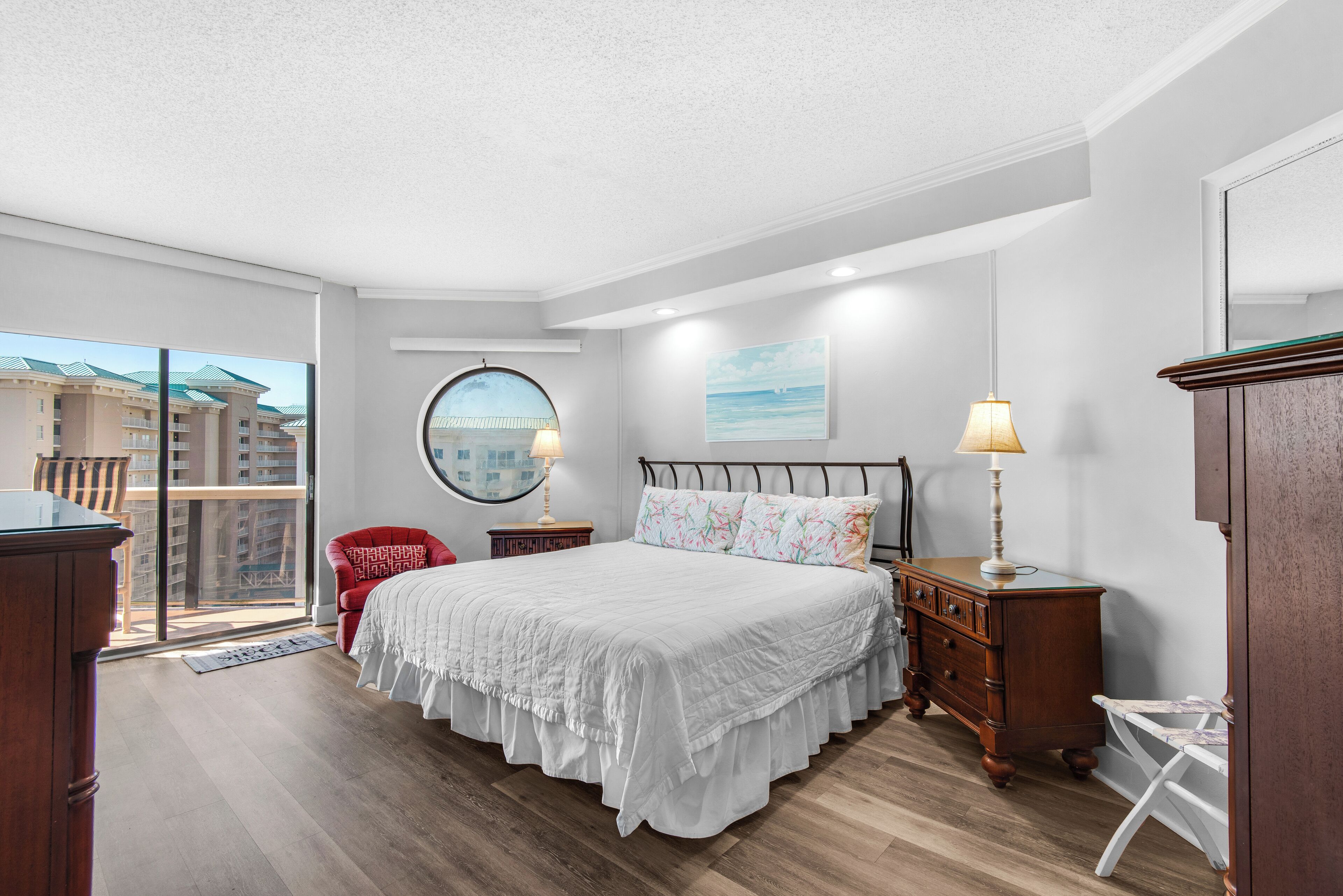 Une chambre à coucher avec un grand lit, un fauteuil rouge, une commode en bois et une vue sur les immeubles par une grande fenêtre.