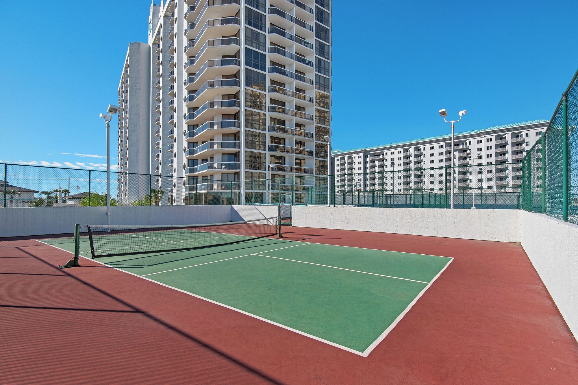 Un court de tennis en plein air, doté d’un filet et entouré d’une clôture, situé à côté d’un grand immeuble d’habitation.