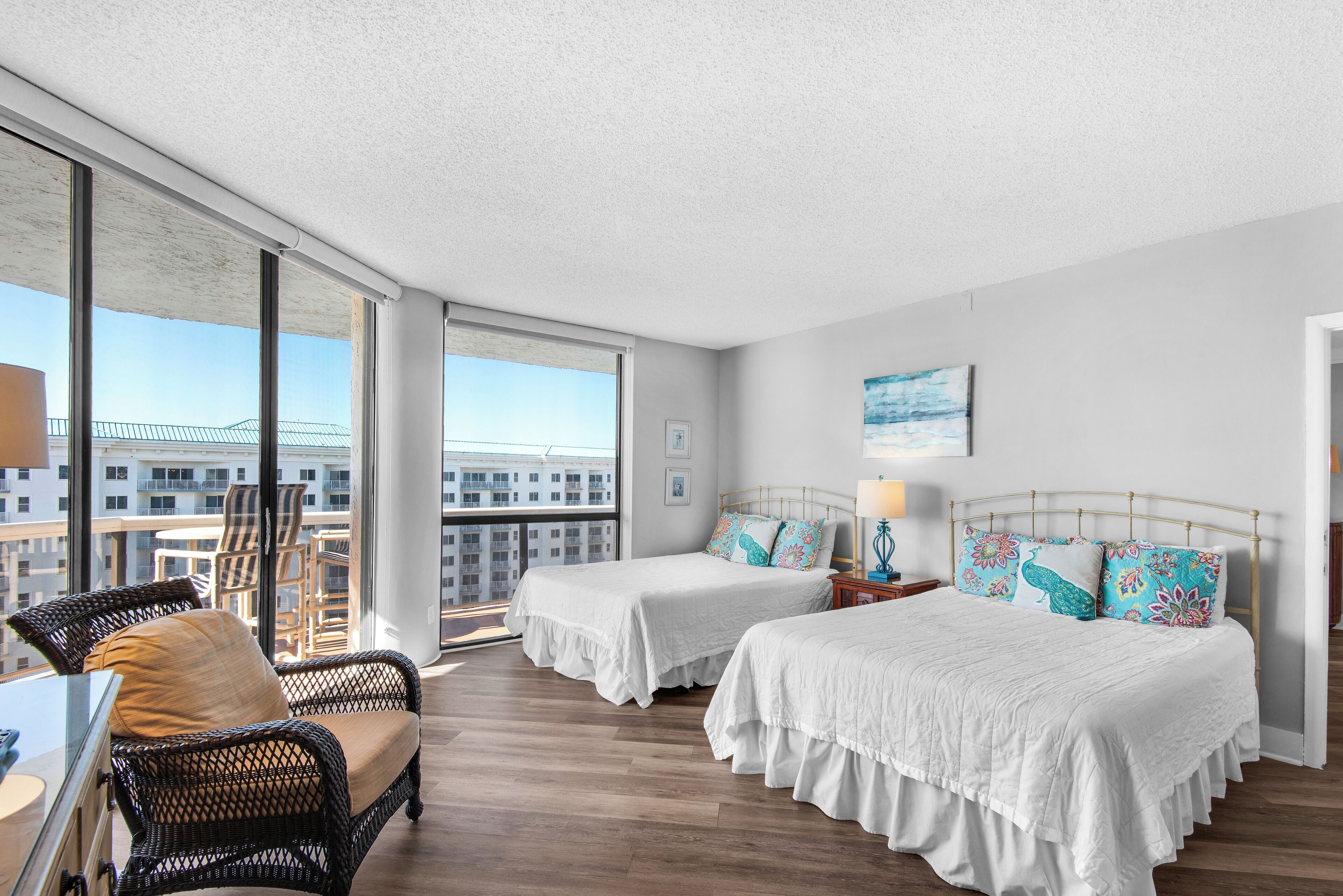 A hotel room with two beds, a balcony with a view of the city, wicker furniture, and a large window.