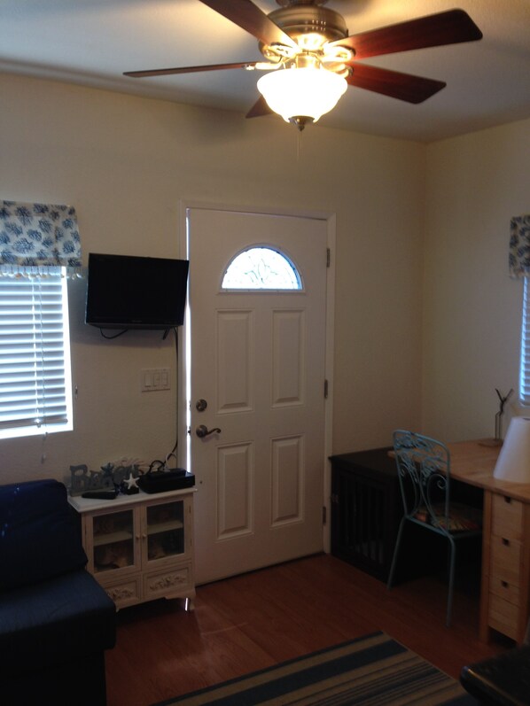 A room with a ceiling fan, a door with a window, a television mounted on the wall, a small cabinet, a chair, and a desk.