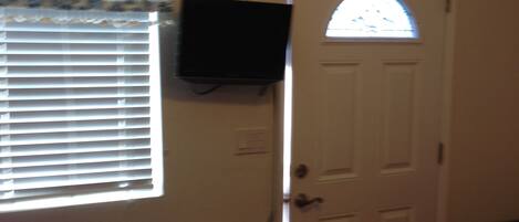 A room with a white door, a television mounted on the wall, a window with blinds, and a small white cabinet.