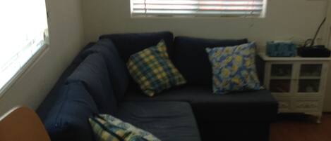 A living room with a dark blue sofa, two patterned pillows, a wooden chair, a small white cabinet, and a window with blinds.