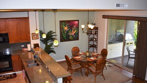 A modern kitchen with granite countertops, wooden cabinets, and a dining area with a glass table and wicker chairs. - Welcome to 1604, Our Little Piece of Paradise! (Waimea)