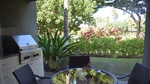 An outdoor patio with a grill, wicker chairs, a glass table, and a view of a garden. - Welcome to 1604, Our Little Piece of Paradise! (Waimea)