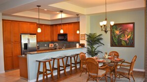 A modern kitchen with wooden cabinets, a stainless steel refrigerator, a microwave, and a bar area with stools. - Welcome to 1604, Our Little Piece of Paradise! (Waimea)