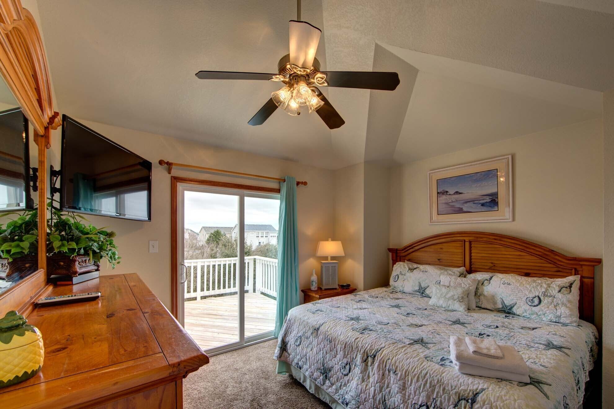 A bedroom with a bed, a ceiling fan, a painting, and a view of the outside.
