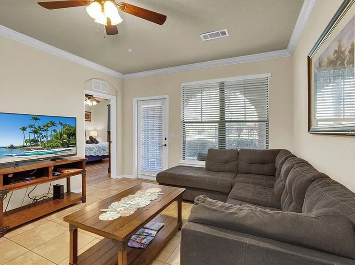 A living room with a television, sofa, coffee table, ceiling fan, and large windows.