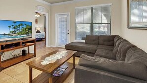 Ein Wohnzimmer mit Fernseher, Sofa, Couchtisch, Deckenventilator und großen Fenstern.